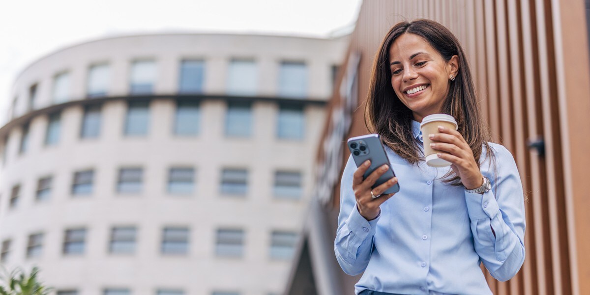 professional woman smiling, standing outside of building, holding cup of coffee and cellphone